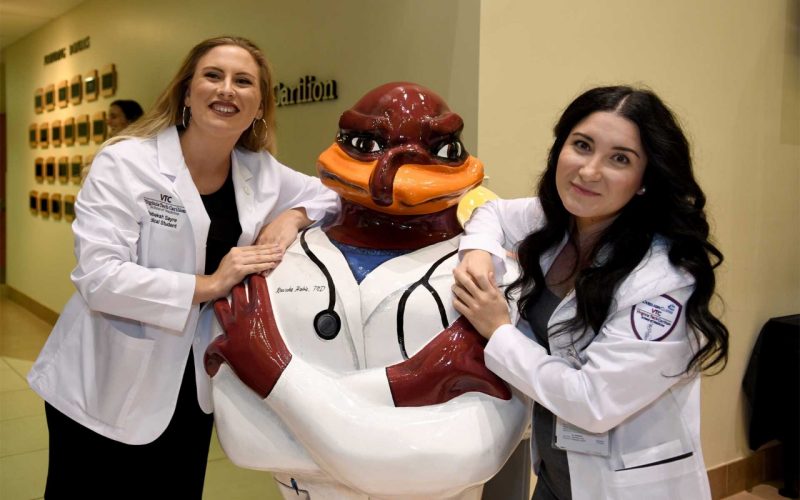 Two female students from the class of 2023 posing with the Roanoke Hokie MD statue at their white coat ceremony