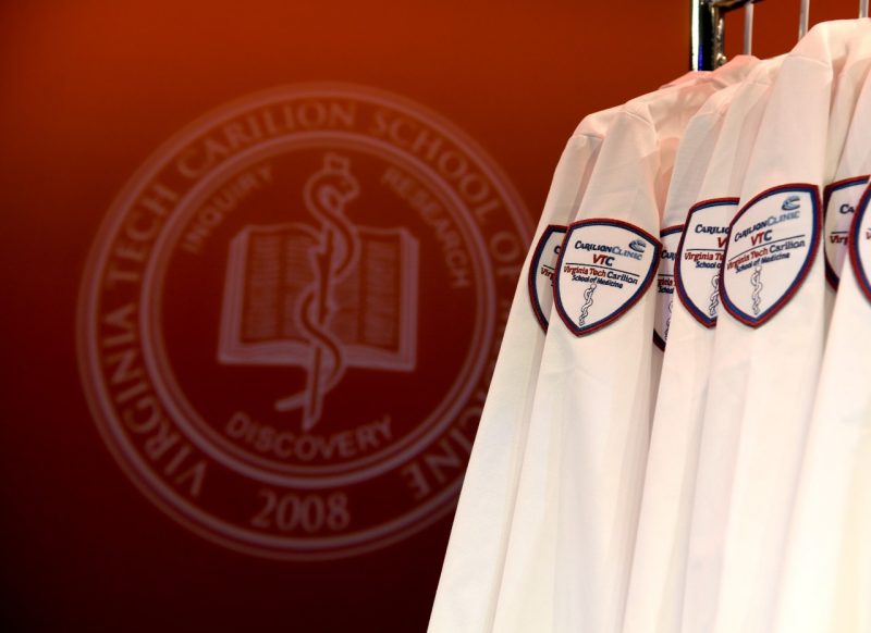 White physician coats containing the Virginia Tech Carilion School of Medicine patch. A larger Virginia Tech Carilion School of Medicine seal is in the background.