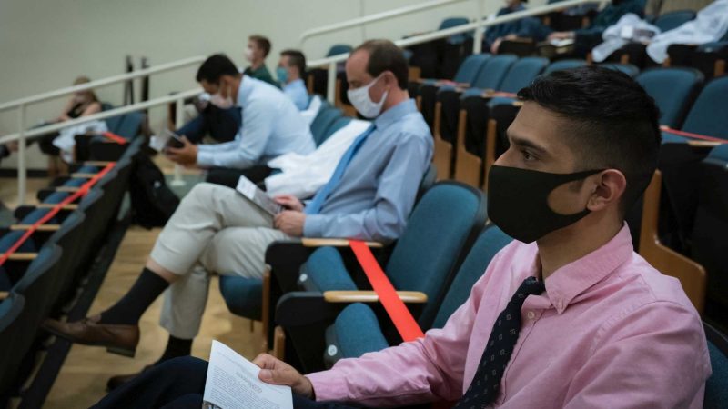 close up of a row of masked students, physically spaced, in the auditorium. Red tape marks where seats are not available. 