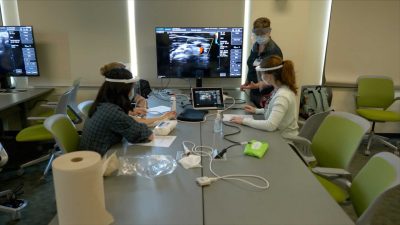 Four students, each wearing masks and faceshields, are seated around a table, looking at a large screen with an ultrasound image