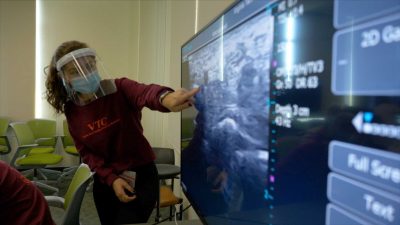 A student, wearing a mask and face shield, points at an ultrasound image on a large screen