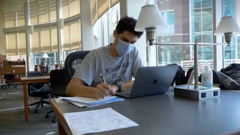 A student, wearing a mask, is taking notes while viewing his laptop.