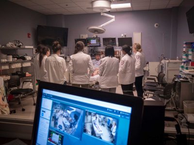 Foreground a screen with footage from two cameras. In the background we see seven medical students talking to a standardized patient in a simulated exam room.