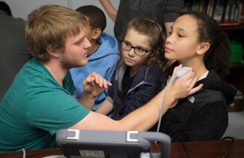 Class of 2019 student Spencer Lovegrove demonstrates a portable ultrasound to two young girls by holding the ultrasound to one of the girls' neck