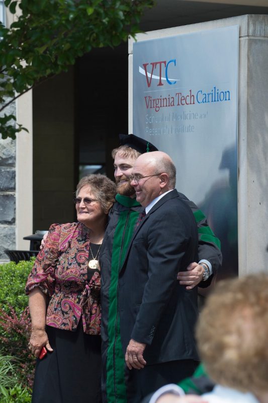 Dr. Daniel Fish stands with his parents in front of the sign at the Virginia Tech Carilion School of Medicine after graduation exercises for the Class of 2016.