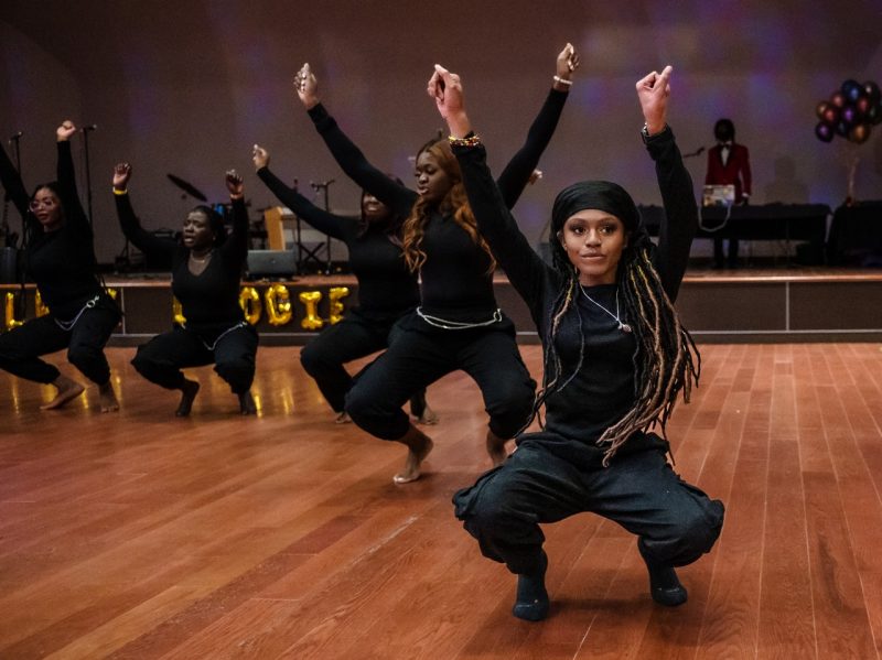 Students of the Wahala Dance group perform at the 2022 Black Excellence Gala.  One female dancer dressed all in black is in focus with other members of her dance group fanned out behind her.