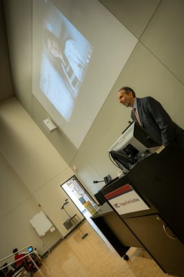 Man speaking from behind a podium with photo of woman projected on high screen