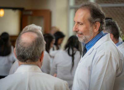 Dean Lee Learman speaking with people at a reception with medical students wearing white coats in background.