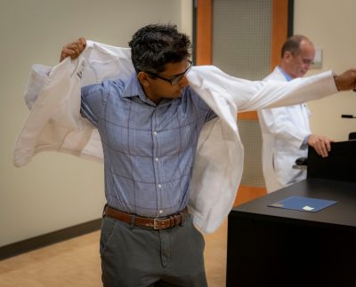 Medical student Varun Kavuru puts on his white coat at the front opf an auditorium. Senior Dean Aubrey Knight stands at podium in background.