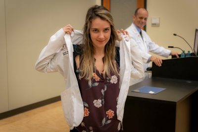 Medical Student Sarah Anderson putting on her white coat in auditorium with Senior Dean Aubrey Knight at podium in background