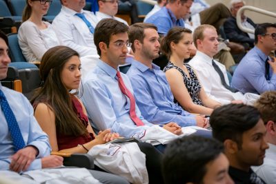 Medical students, holding white coats in their laps, seated in auditorium.