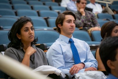 Male and femaie medical students sit side-by-side in auditorium with more students seated in background.