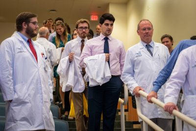 Medical students, carrying white coats over their arms, and physicians processing single file. 