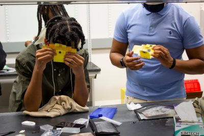 Two male students are looking through their yellow and orange foldscopes.