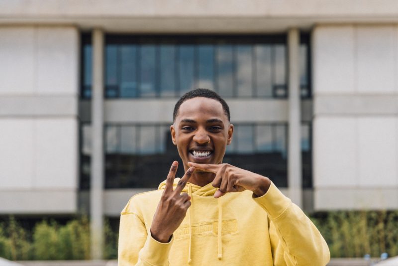 A Black man in a yellow hoodie makes a VT symbol with his hands.