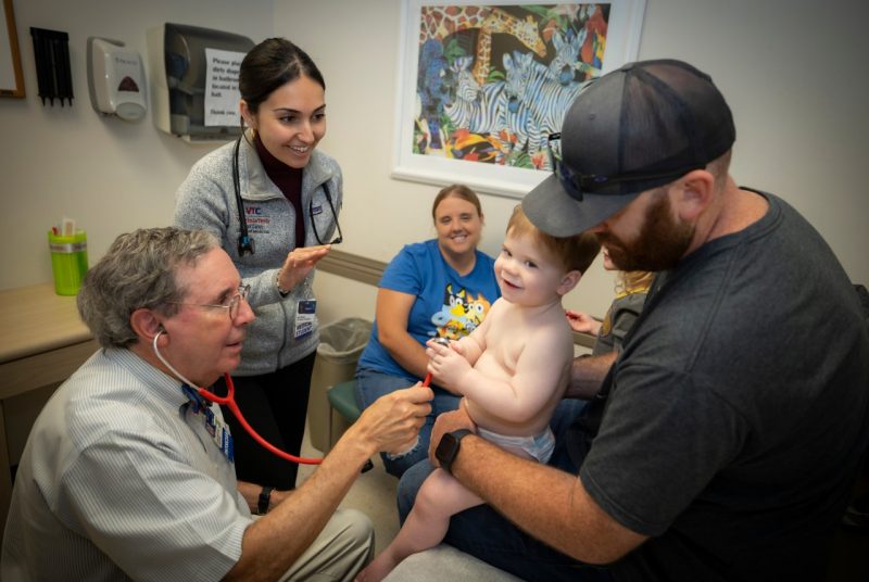 Dan with stethoscope listens to toddler's chest as female medical student watches. The toddler is being held by his father. His mother is sitting in the background./