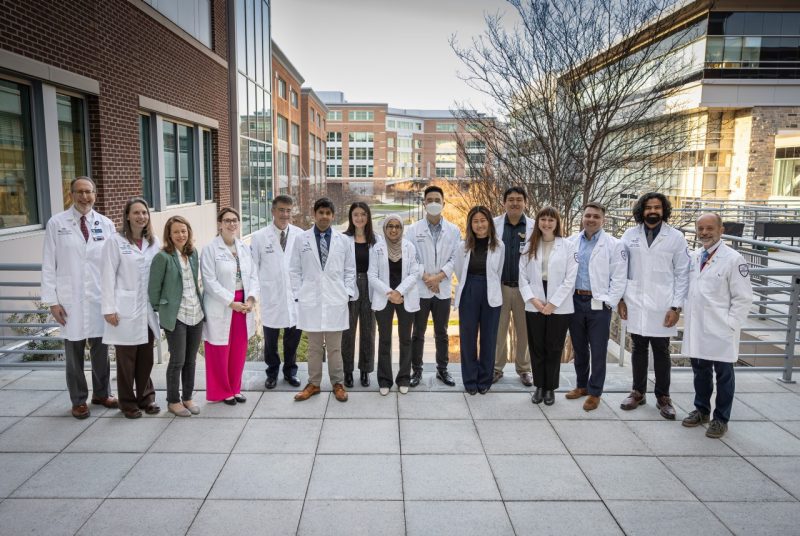 Fourteen individuals, most wearing white doctor's coats are aligned side-by-side outdoors.