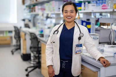 A medical student with a white lab coat and a stethoscope stands in a lab setting.