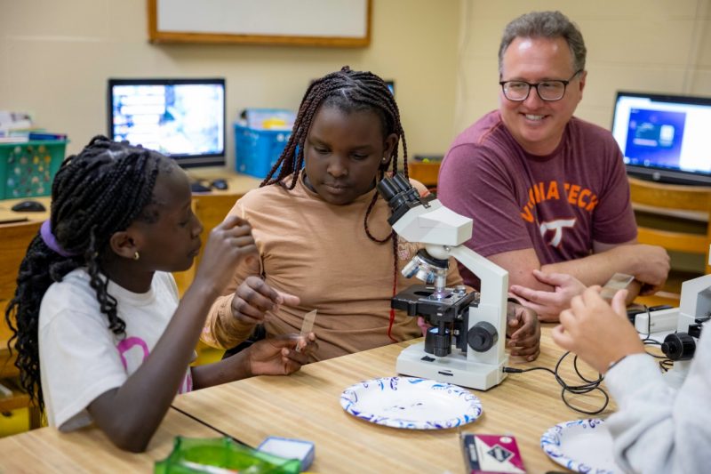 Two children work with a microscope while an adult man looks on. 