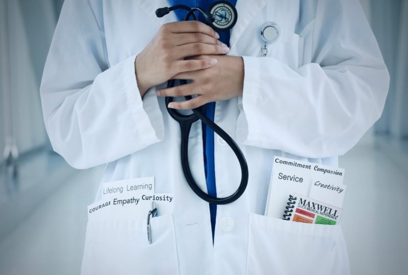 photo of someone in a whitecoat from the shoulders down to the waist. the person is holding a stethoscope. inside the pockets are the words: lifelong learning, courage, empathy, curiosity; commitment, compassion, service, creativity, and a maxwell book