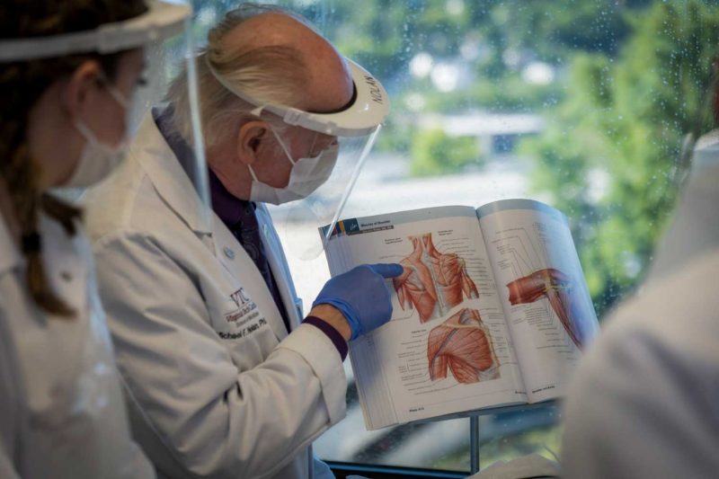 instructor wearing personal protective equipment such as face shield mask and glove, pointing at an image of a shoulder in a book. Beside him stands a student also wearing PPE.