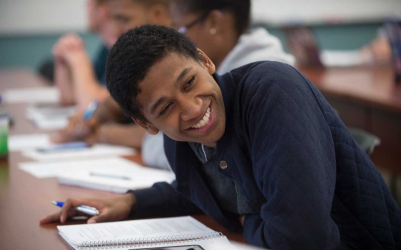 Young Black male leaning over a desk looking off camera with a big smile. 