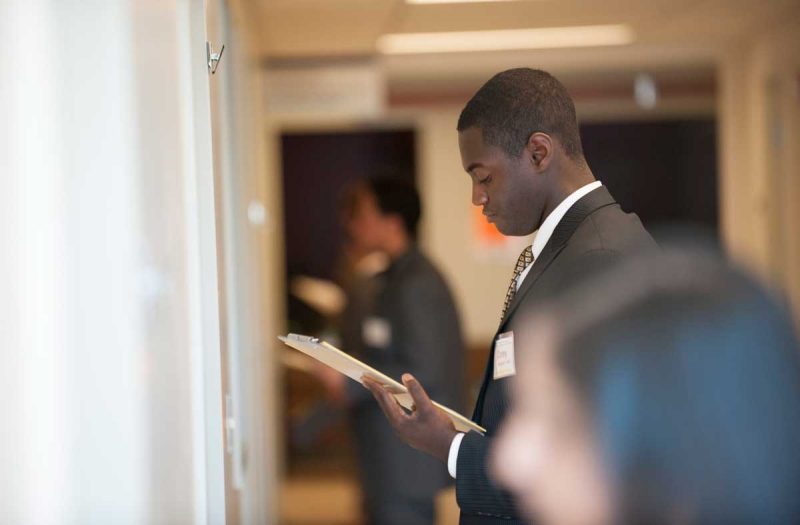 focus is on an african american male wearing a suit holding a clip board in a hallway. three other students are blurred