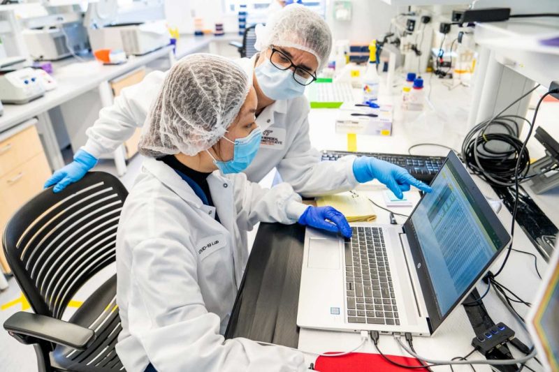 Two researchers wearing hair nets and masks in the lab reviewing data on a laptop. 