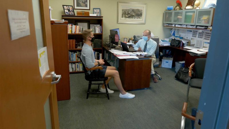 View of a student and the senior dean for student affairs interacting in his office. Both are wearing masks. 