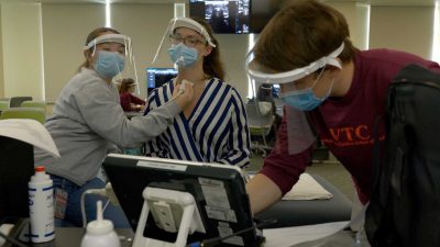three students, wearing masks and face shields. Student one holds an ultrasound wand to the neck of student two, while student three reaches toward the tablet