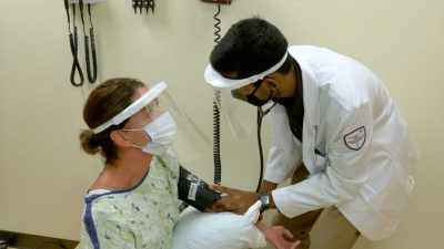 Seated standardized patient looks up at standing student while he takes her blood pressure. Both are wearing masks and face shields.