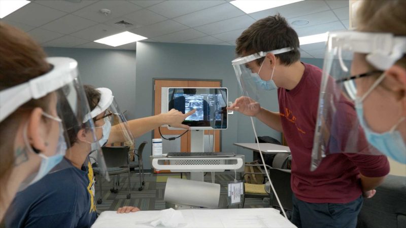 Four students, each wearing masks and face shields, working with an ultrasound machine.