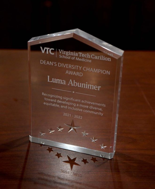 The Dean's Diversity Champion Award, a clear crystal award with stars and inscribed to Luma Abunimer, stands on a wood table.
