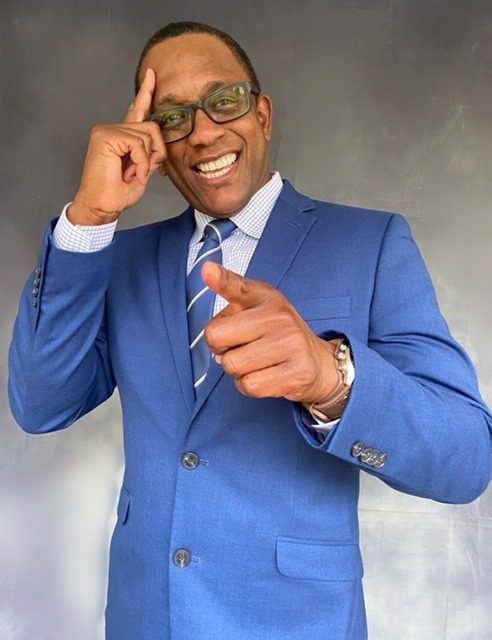 Alexander Scott, african american male in a blue suit and wearing glasses, saluting to the camera with a big smile