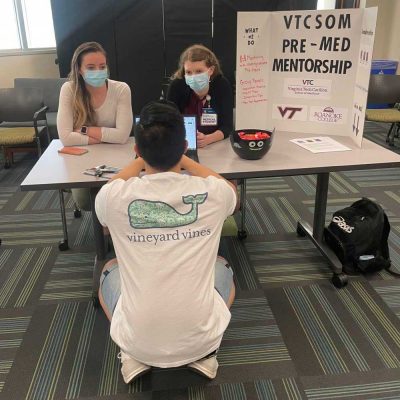 Two female students (masked) sitting at a table with a VTCSOM Pre Med Mentorship board. A male student crouched down in front of them with a vineyard vines t-shirt.