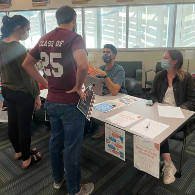 two students stand in front of a table with dermatology interest group flyers. Two student sitting at the table representing that group. 