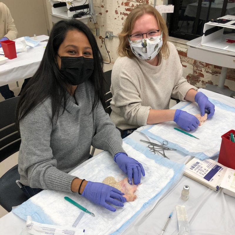 Two students smiling at the camera. They are wearing masks and gloves. In their gloved hands they are holding practice prosthetic body parts. On the table they have instruments including scalpels and scissors. 