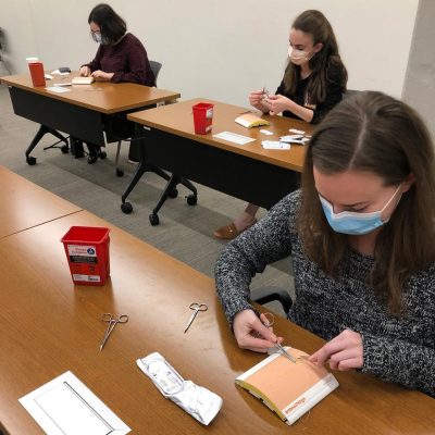 Suture and biopsy workshop. One student in the foreground using tweezers and scissors on a practice canvas. Two students in the background practicing sutures also.